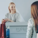 Kasir menyerahkan tas belanja kepada pelanggan di toko, menggambarkan customer retention yang baik